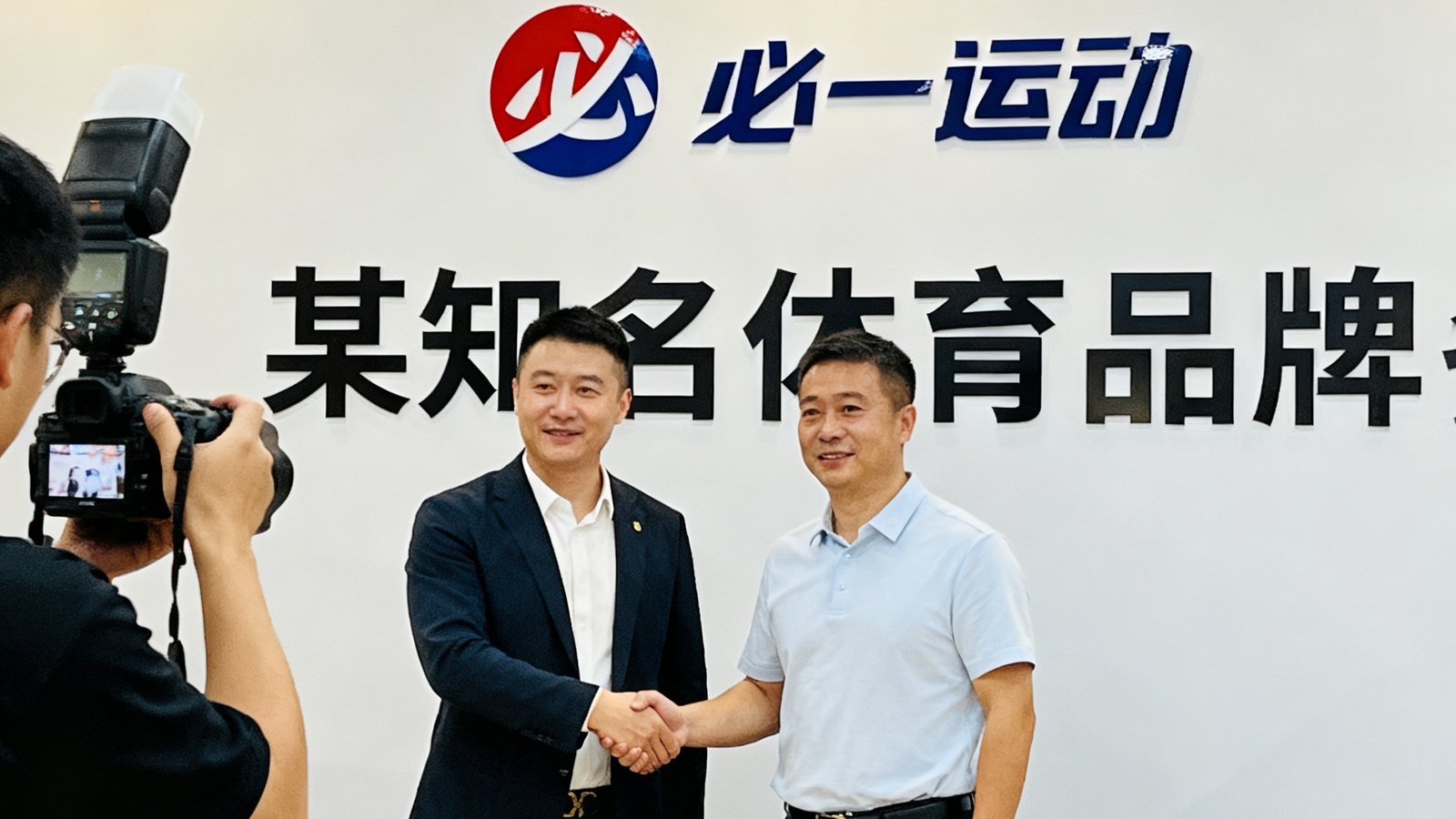 必一运动CEO与某知名体育品牌负责人在签约仪式上握手，双方代表站在印有两家公司logo的背景板前，摄影师正在拍照记录