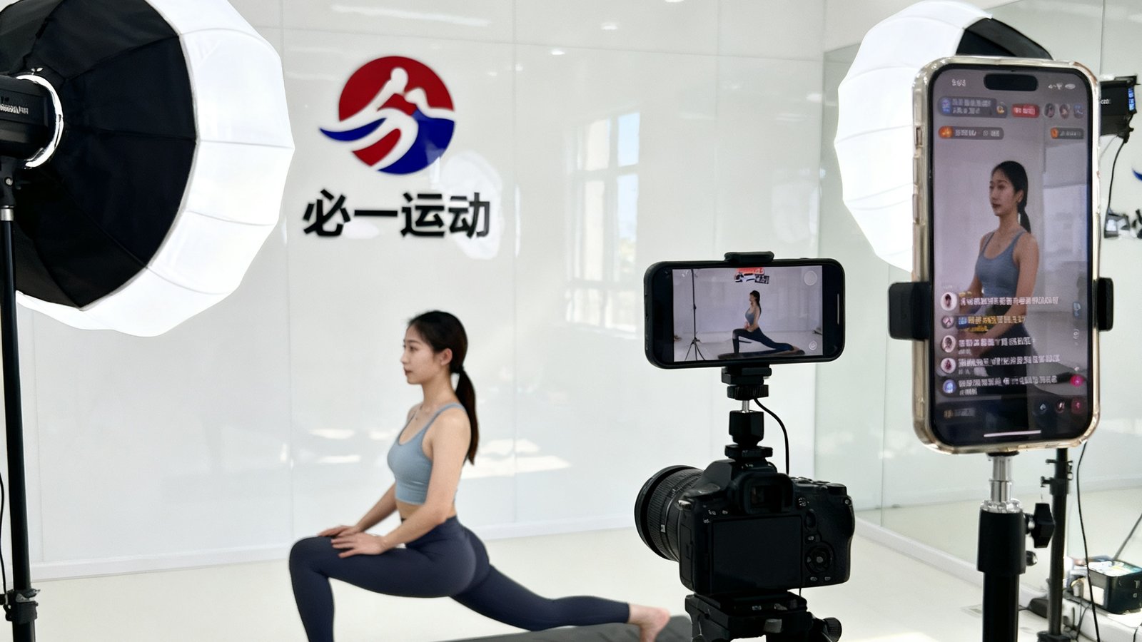 明亮整洁的直播间内，一位女性教练正对着镜头示范瑜伽动作，面前有专业补光灯和摄像机，背景墙上挂着必一运动品牌logo，手机支架上同步显示着学员的实时弹幕互动